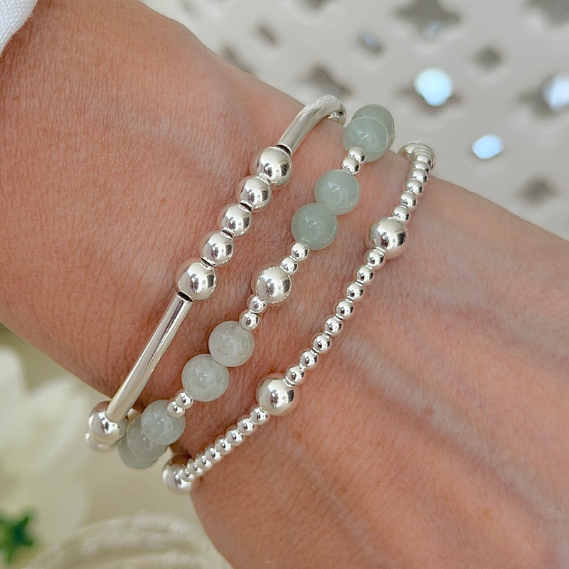 Close-up of a hand wearing two silver beaded bracelets and a third stacking bracelet with green beads mixed with silver, on a blurred background.
