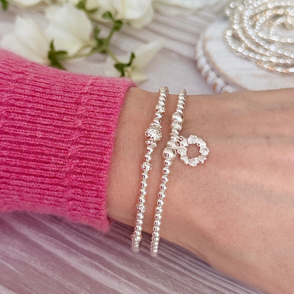 Silver beaded stacking bracelets on a wrist, one featuring a sterling silver wreath shaped charm, with a pink sleeve and floral background