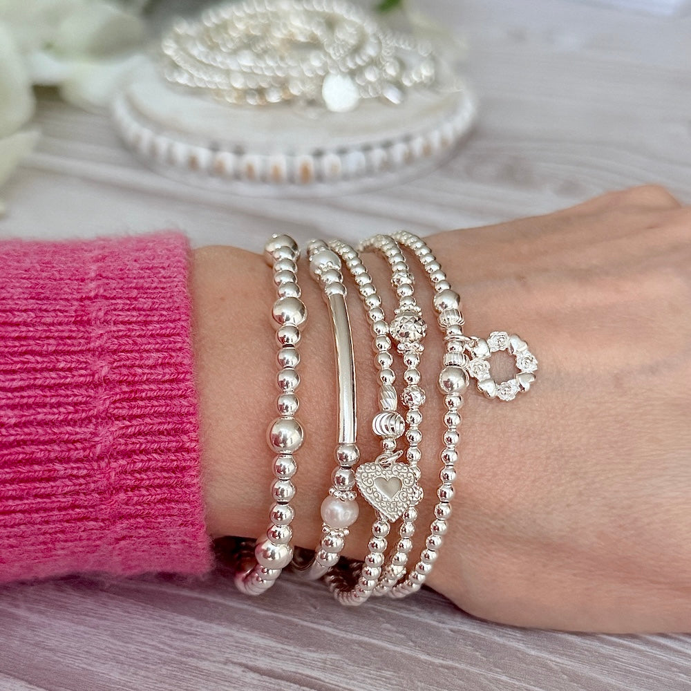 A close up of a lady's wrist wearing a stack of silver stacking bracelets on a wrist with a pink sleeve and blurred background. One of the bracelets features a silver wreath charm and another a small heart charm.