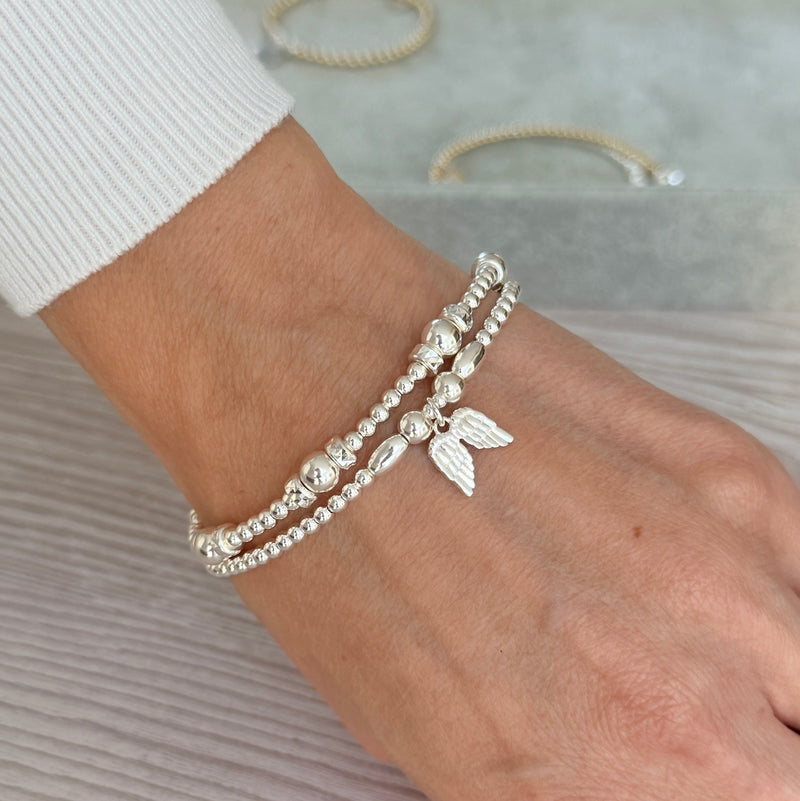 A close-up of a person's wrist wearing two delicate silver beaded bracelets, one featuring a small winged charm, with a white ribbed sleeve visible.