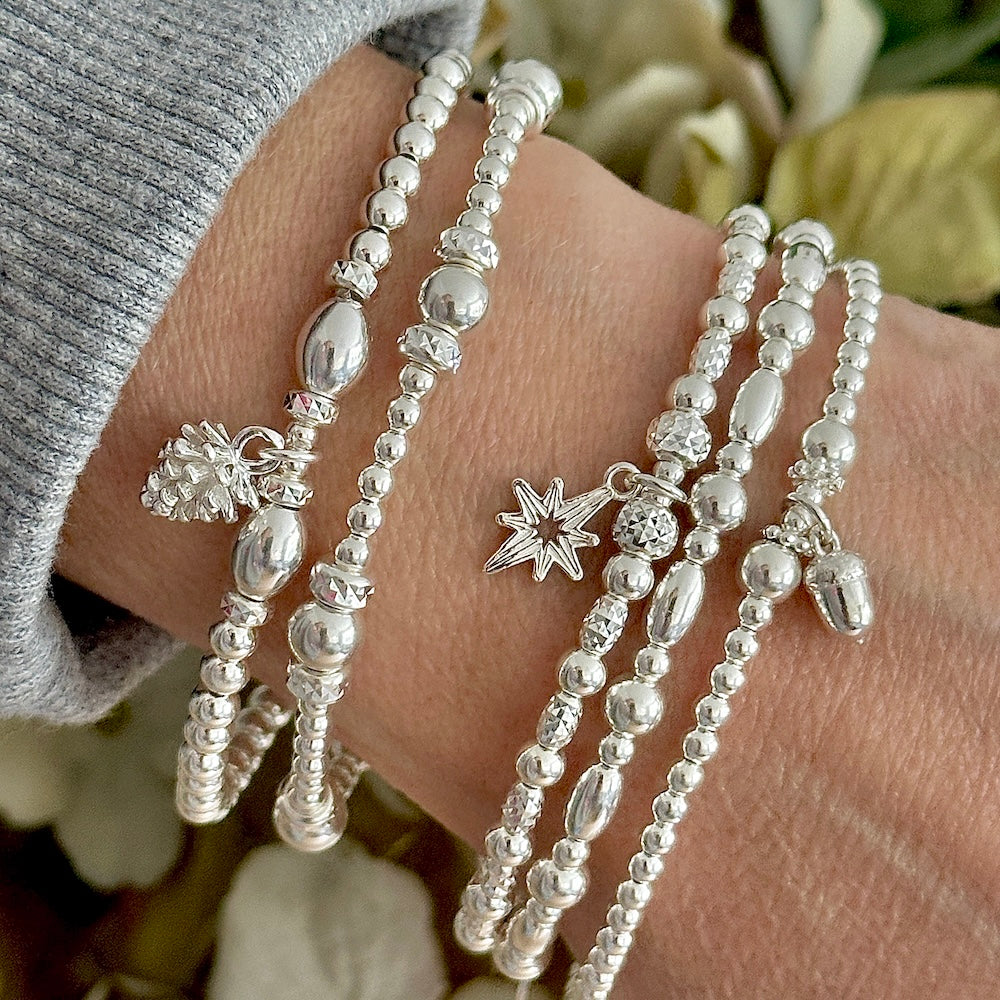 Silver beaded bracelets with star charm, pine cone charm and tiny acorn charm, on a wrist against a floral background