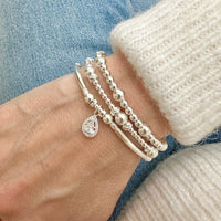 Silver beaded bracelet with a teardrop charm, with two other silver beaded stacking bracelets, on a wrist against a denim background