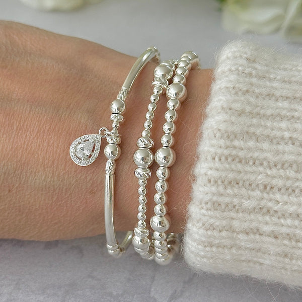 Silver bracelet bracelet stack with one bracelet featuring a sparkly tear drop charm, on a wrist against a neutral background