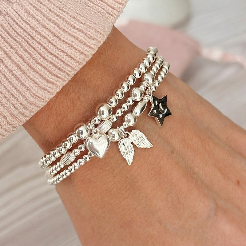A close-up of a person's wrist wearing three stacked sterling silver beaded bracelets, featuring an angel wings charm, a puffed heart charm and a star charm, styled with a pink knitted jumper.
