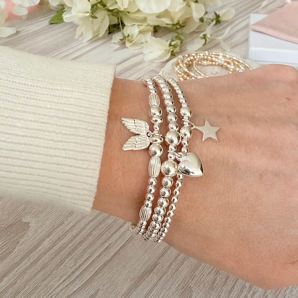 A close up shot of a lady's wrist wearing 3 Bella Jane silver beaded stacking bracelets with a heart, star and angel wings charms, with a cream coloured jumper sleeve, against a neutral background with white flowers.