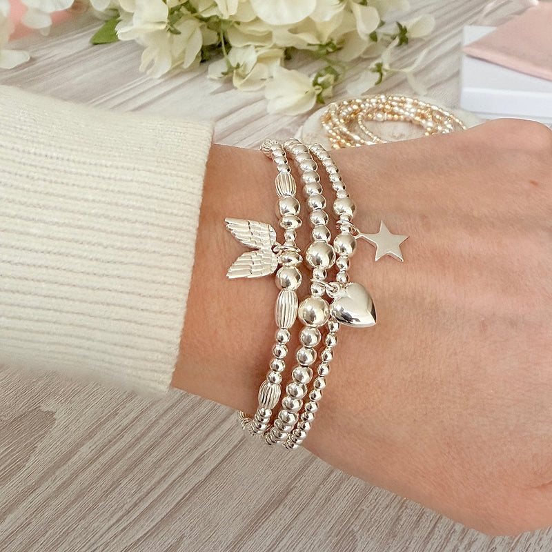 A close up shot of a lady's wrist wearing 3 Bella Jane silver beaded stacking bracelets with a heart, star and angel wings charms, with a cream coloured jumper sleeve, against a neutral background with white flowers.