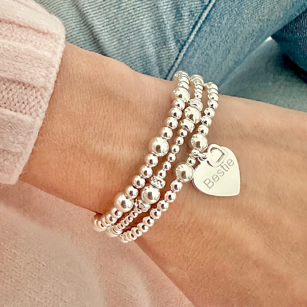 Three silver beaded bracelets with a heart-shaped charm on one of the bracelets, engraved with the word 'Bestie' -  on a wrist against a denim jeans background.