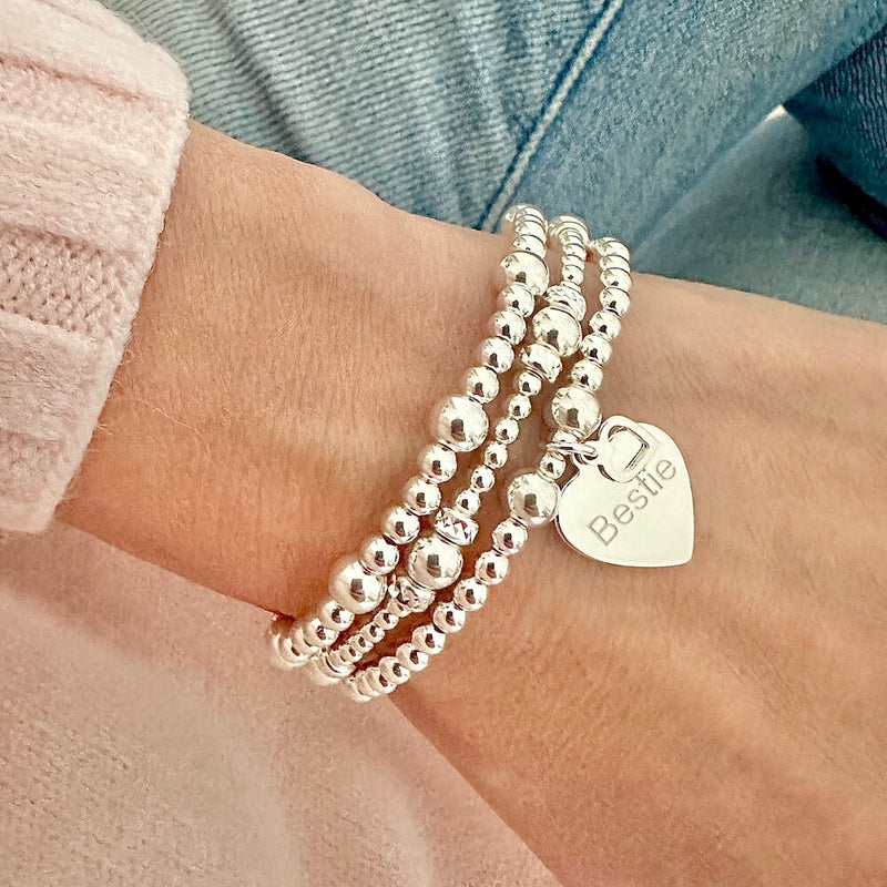 Three silver beaded bracelets with a heart-shaped charm on one of the bracelets, engraved with the word 'Bestie' -  on a wrist against a denim jeans background.
