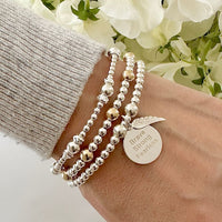 Silver beaded bracelet with the words Brave, strong, fearless engraved on a silver disc charm, with a small angel wing charm, worn on a wrist with two other beaded bracelets, against a floral background.