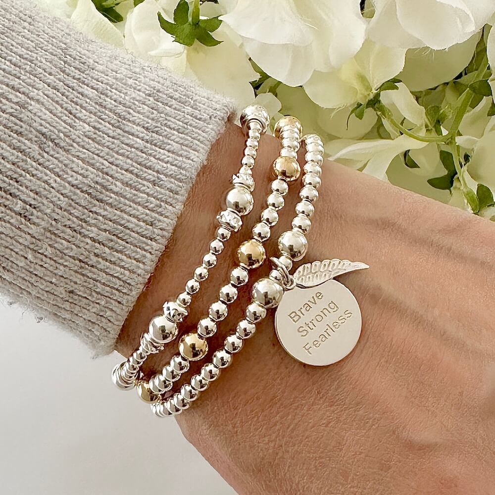 Silver beaded bracelet with the words Brave, strong, fearless engraved on a silver disc charm, with a small angel wing charm, worn on a wrist with two other beaded bracelets, against a floral background.