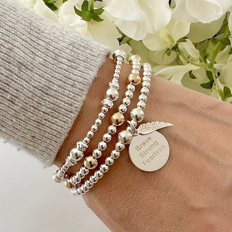 Silver beaded bracelet with the words Brave, strong, fearless engraved on a silver disc charm, with a small angel wing charm, worn on a wrist with two other beaded bracelets, against a floral background.