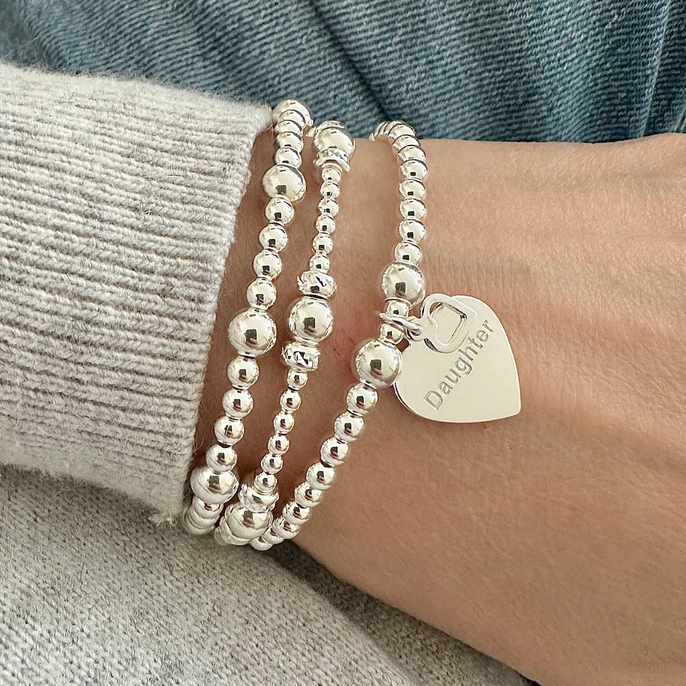 Silver beaded bracelets on a wrist with one bracelet featuring a heart-shaped charm with the word Daughter engraved on it with a tiny heart charm.