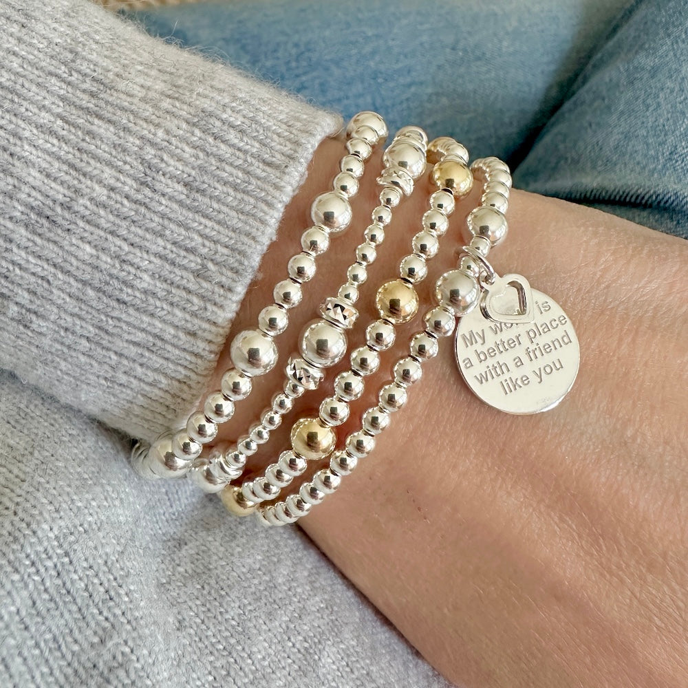 A wrist wearing a stack of 4 silver and silver/gold beaded bracelets on a wrist with one bracelet featuring a round silver charm with tiny heart and the words 'My world is a better place with a friend like you' engraved on the front.