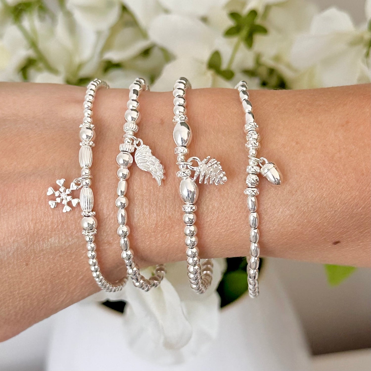 A wrist wearing 4 sterling silver beaded bracelets with a snowflake, angel wing, pine cone and acorn charms