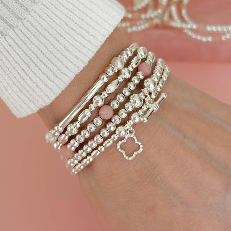 Stack of silver beaded bracelets on a hand, with one bracelet featuring pink opal beads with the silver, against a soft pink background