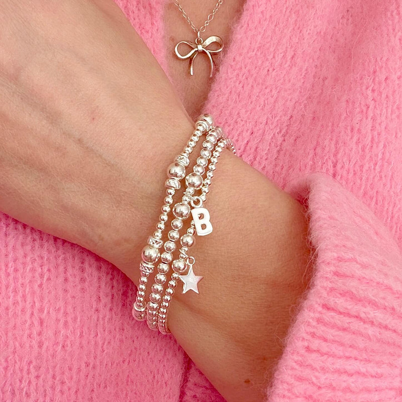 Silver beaded bracelets with initial 'B' and star charms on a wrist against pink sweater, with a silver bow necklace
