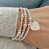 A wrist wearing three silver beaded stacking bracelets, one with a heart-shaped charm with the words 'Love, Luck, Happiness' engraved on the front, with a tiny four-leaf clover charm sitting on top.