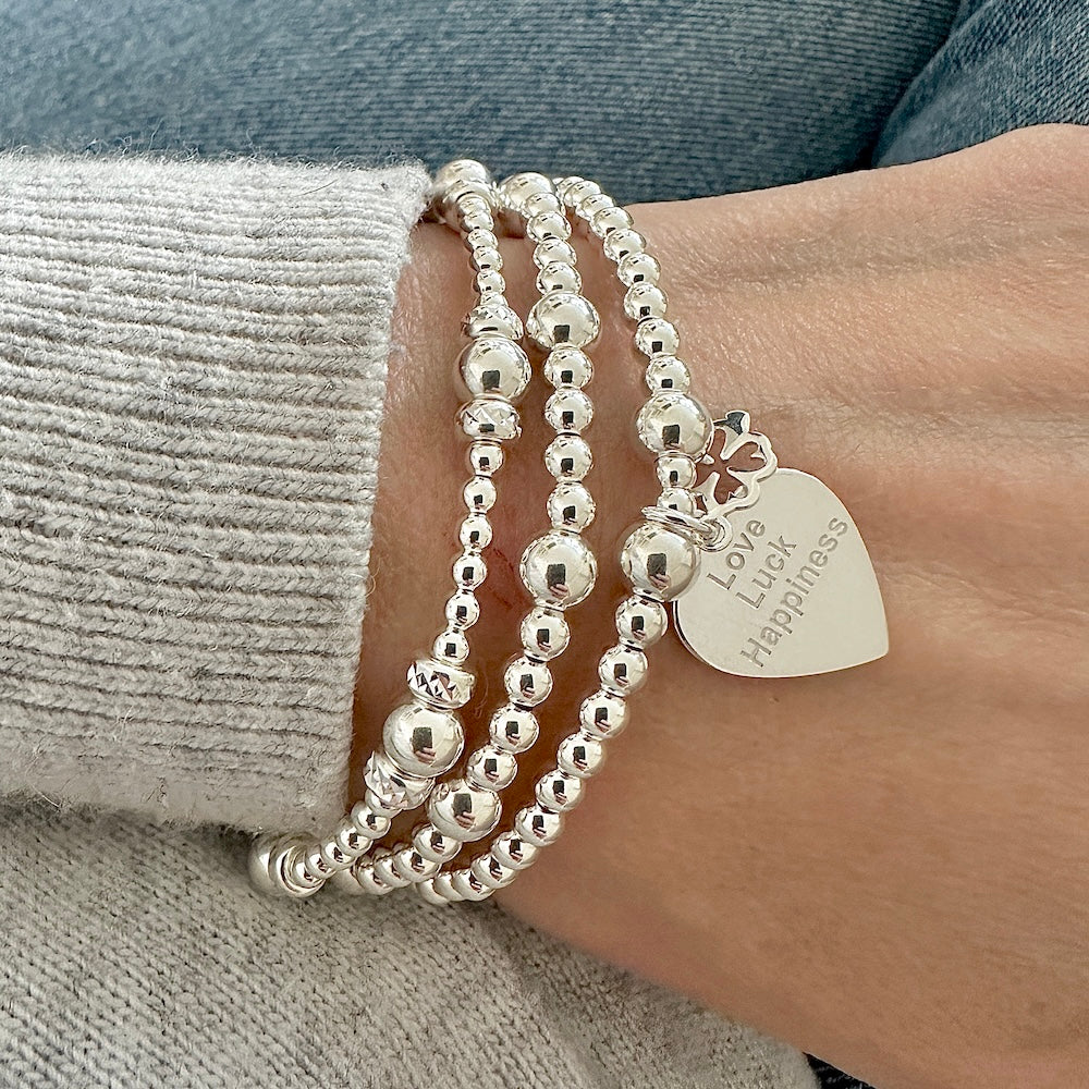 A wrist wearing three silver beaded stacking bracelets, one with a heart-shaped charm with the words 'Love, Luck, Happiness' engraved on the front, with a tiny four-leaf clover charm sitting on top.