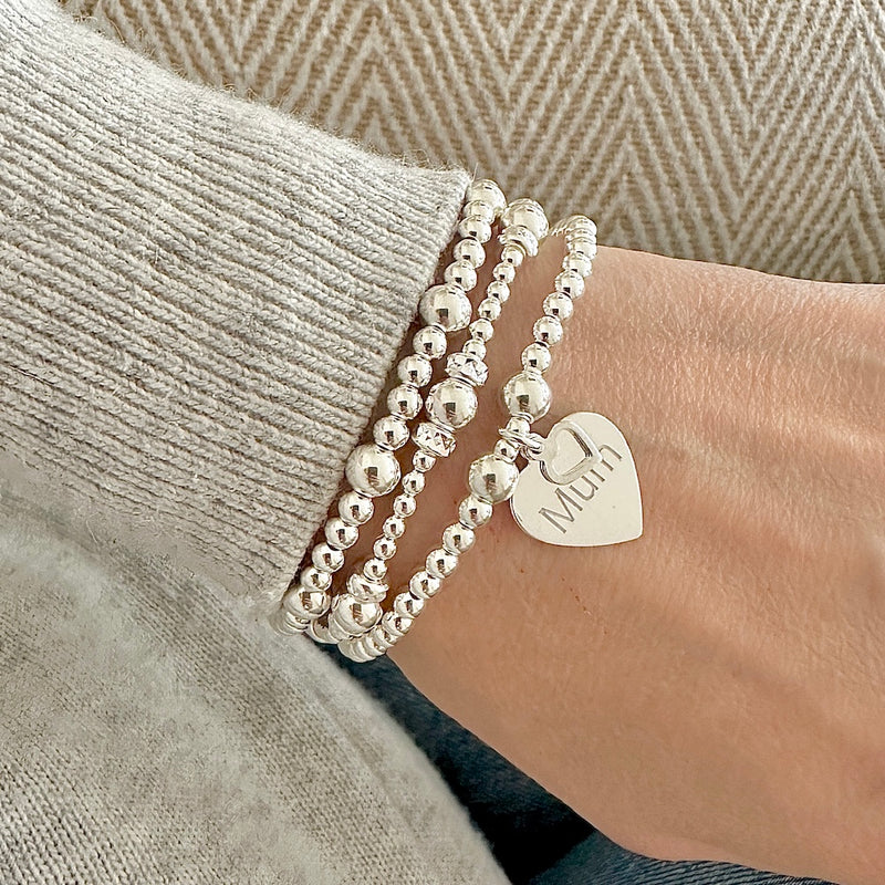 3 silver beaded bracelets with one featuring a heart-shaped charm with the word 'Mum' engraved, on a wrist, wearing a light grey sweater.