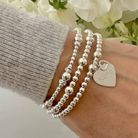 Silver beaded bracelets on a wrist with a heart-shaped charm, featuring the word 'Nana', against a blurred floral background.