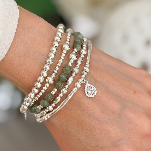 A close up of a lady's wrist wearing silver beaded bracelets with one olive green and silver beaded bracelet, against a neutral background.