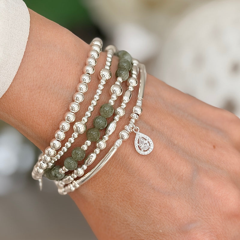 A close up of a lady's wrist wearing silver beaded bracelets with one olive green and silver beaded bracelet, against a neutral background.