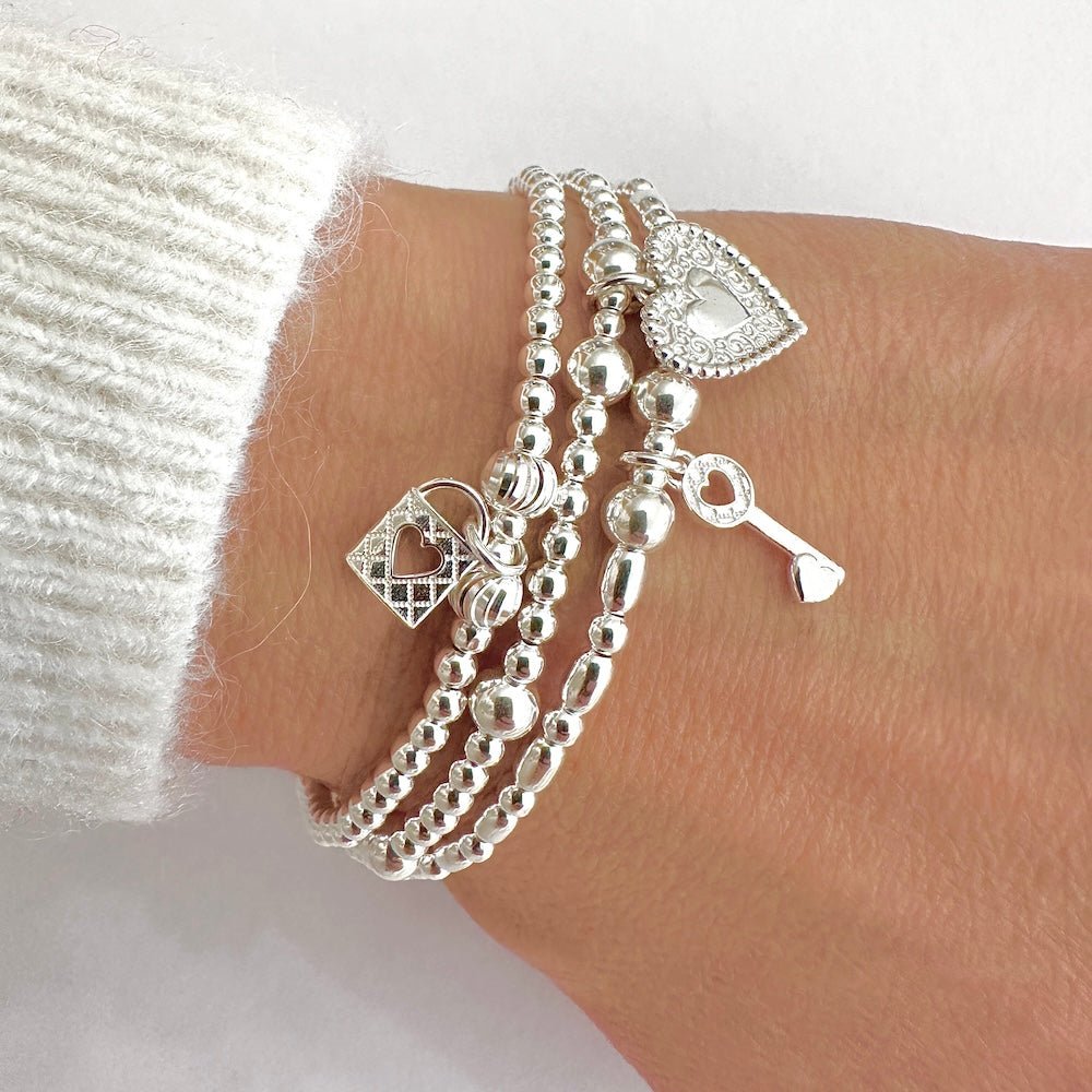A close up of a wrist wearing three silver beaded stacking bracelets, showing a heart padlock, key and heart charms, against a white background