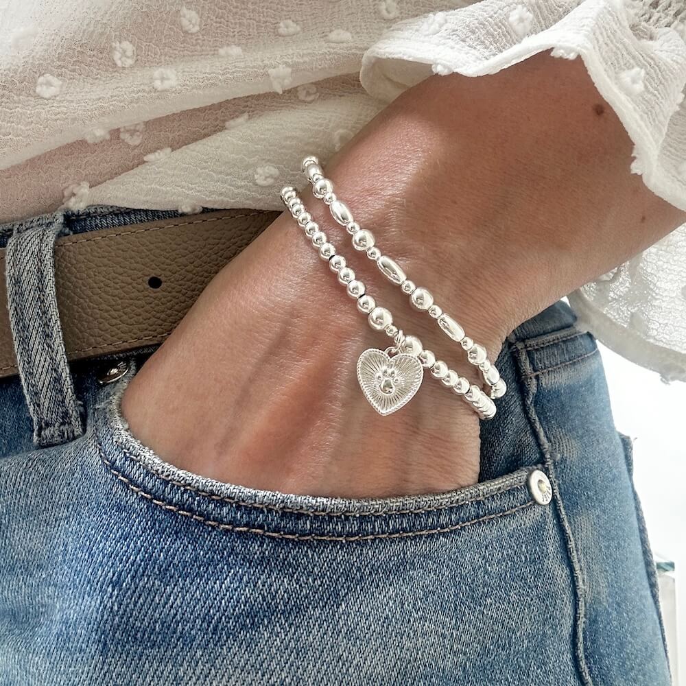 A close up of a woman's wrist wearing a Silver beaded stacking bracelet duo with one bracelet featuring a paw print heart charm, wearing a white blouse and blue jeans.