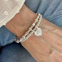 A close up of a woman's wrist wearing a Silver beaded stacking bracelet duo with one bracelet featuring a paw print heart charm, wearing a white blouse and blue jeans.
