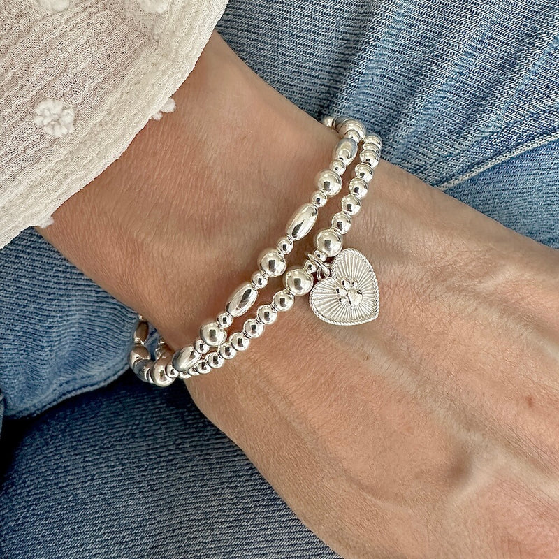 A close up of a woman's wrist wearing a Silver beaded stacking bracelet duo with one bracelet featuring a paw print heart charm, wearing a white blouse and blue jeans.