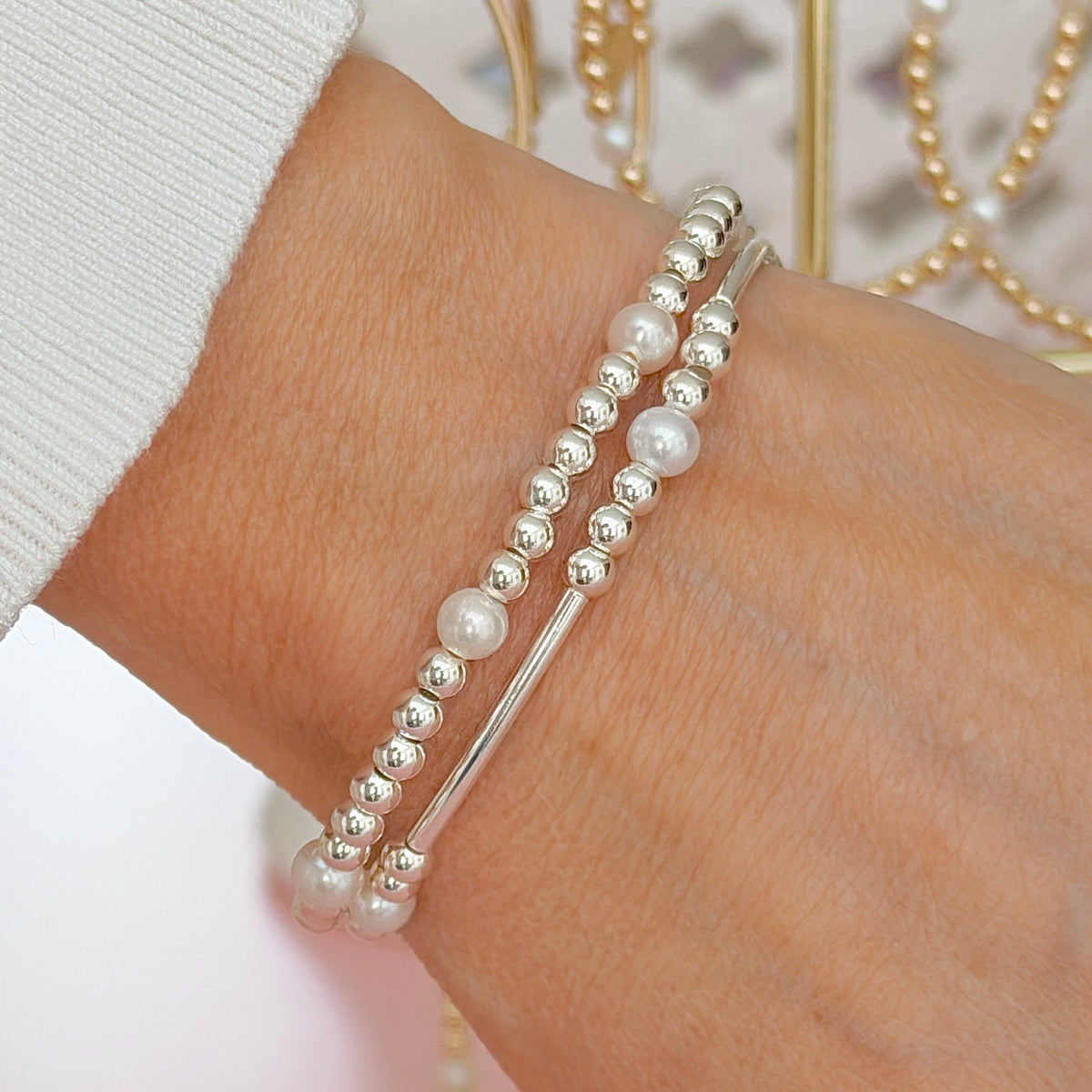 A close-up of a person's wrist wearing two sterling silver and freshwater pearl stacking bracelets with gold bracelets hanging in the background