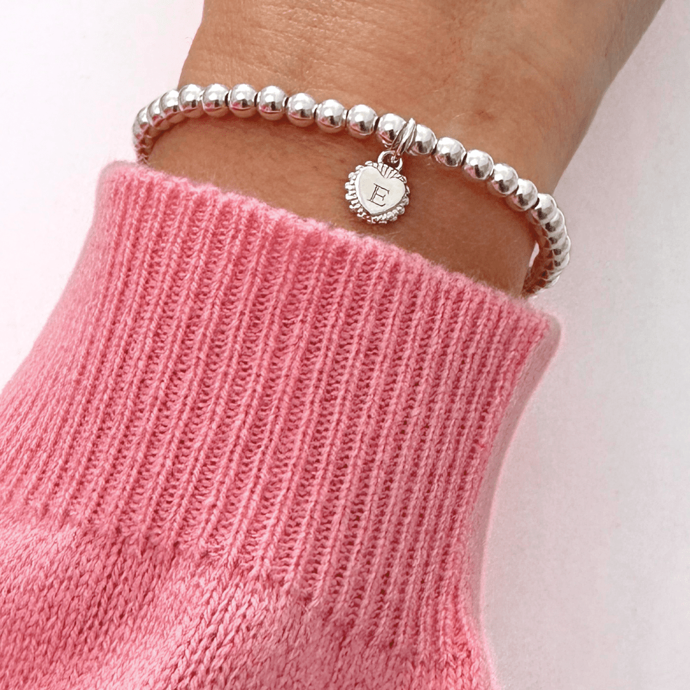 Silver beaded bracelet with a charm engraved with the initial 'E' on a wrist wearing a pink sweater.
