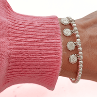 Silver beaded bracelet with 4 heart disc charms with initials engraved on them, on a wrist wearing a pink sleeve.