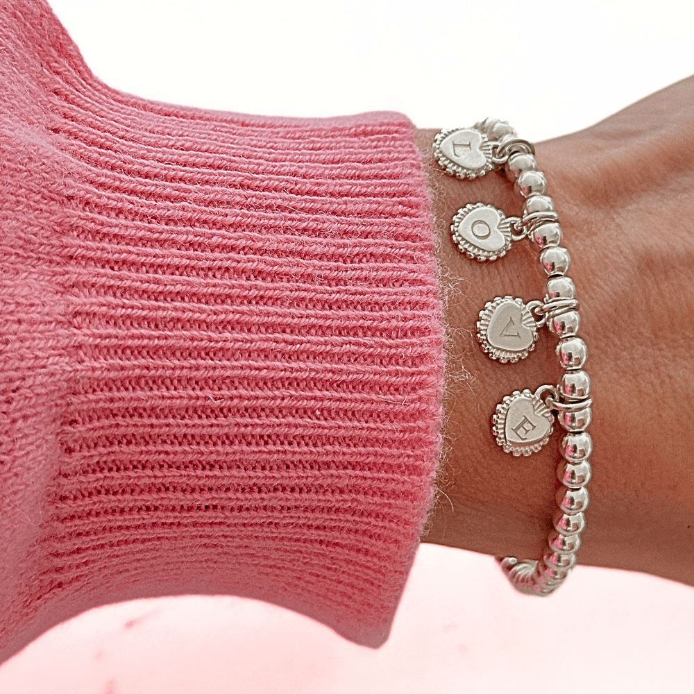Silver beaded bracelet with 4 heart disc charms with initials engraved on them, on a wrist wearing a pink sleeve.