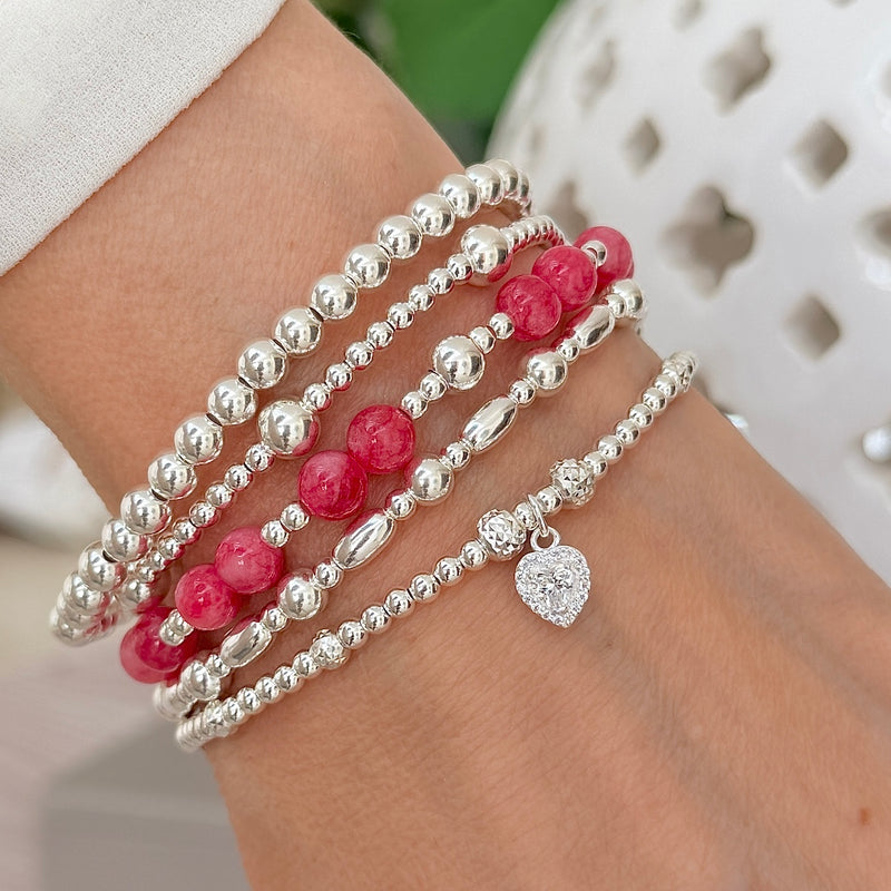 Lady's wrist with four sterling silver beaded bracelets (one with a sparkly heart charm) with one bright raspberry coloured beaded bracelet, featuring silver beads.