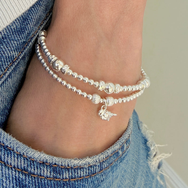 A close-up of a person's wrist wearing two stacked silver beaded bracelets, one with a small robin charm, styled with blue denim jeans.