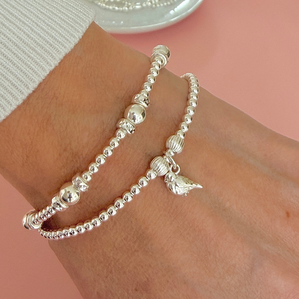Two silver beaded bracelets, one featuring a sterling silver robin charm, worn on a person's wrist against a soft pink background.