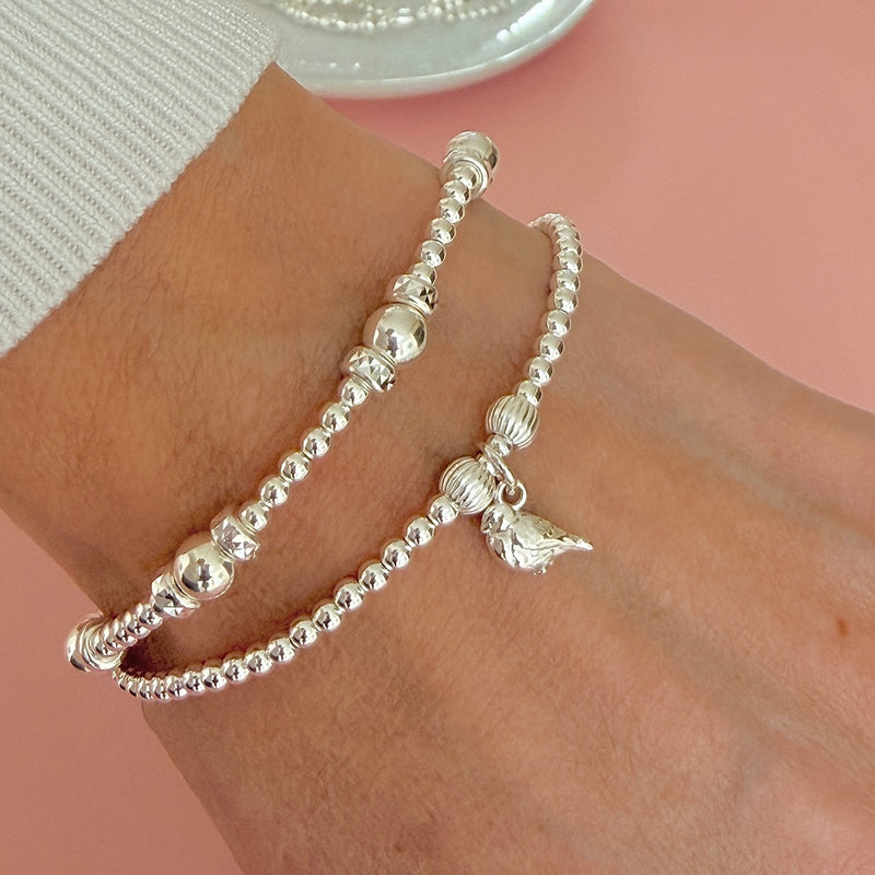 Two silver beaded bracelets, one featuring a sterling silver robin charm, worn on a person's wrist against a soft pink background.