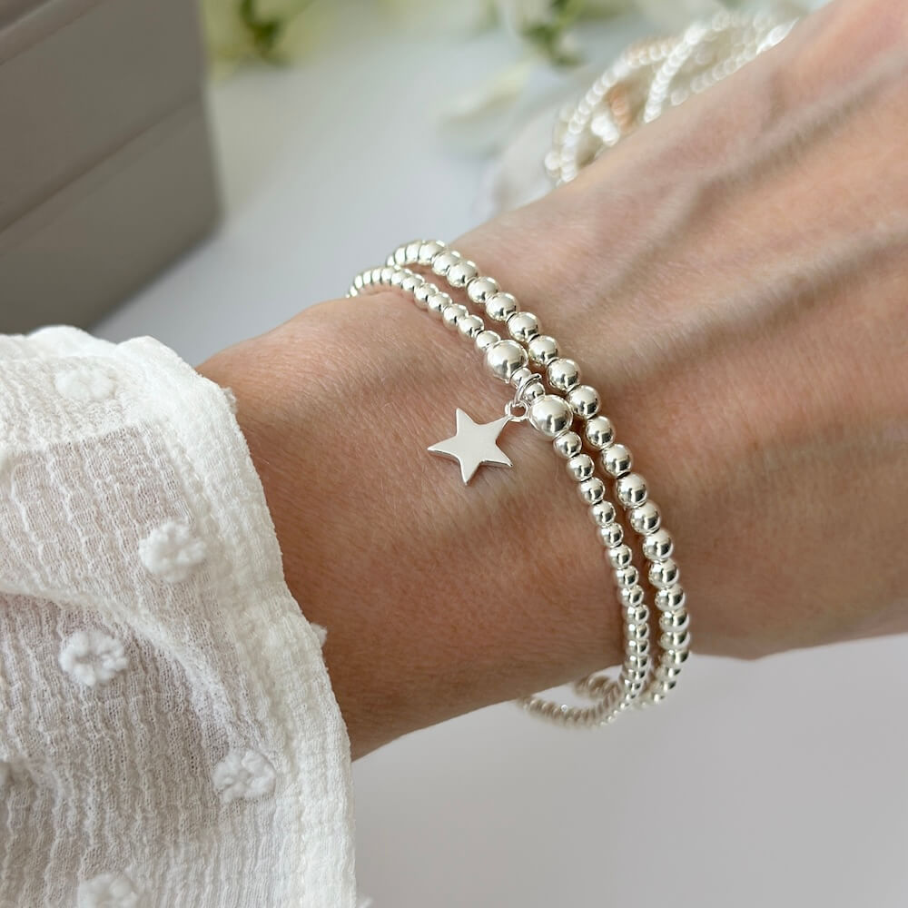 A lady's wrist wearing a silver beaded bracelet duo with a star charm, with a blurred background of flowers.