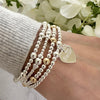 A lady's wrist wearing four beaded bracelets, one featuring a heart shaped charm with the word 'Sister' engraved on it, with white flowers in the background.