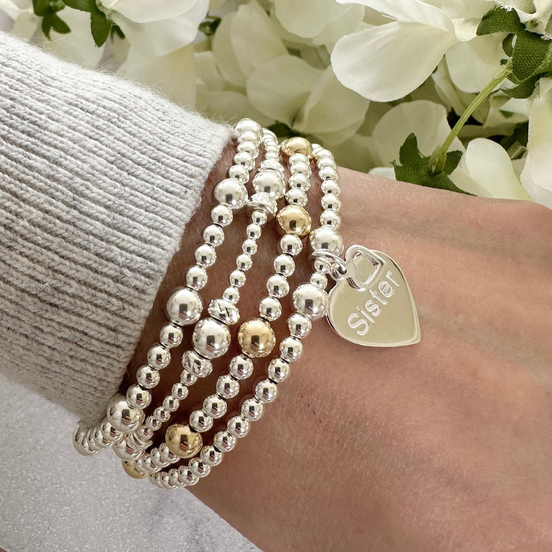 A lady's wrist wearing four beaded bracelets, one featuring a heart shaped charm with the word 'Sister' engraved on it, with white flowers in the background.