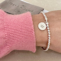 A wrist wearing a silver beaded bracelet with heart disc with the words love you engraved, with a pink jumper sleeve
