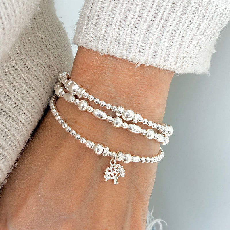 A close up of a person's wrist wearing 3 silver beaded bracelets, with one featuring a small tree charm.