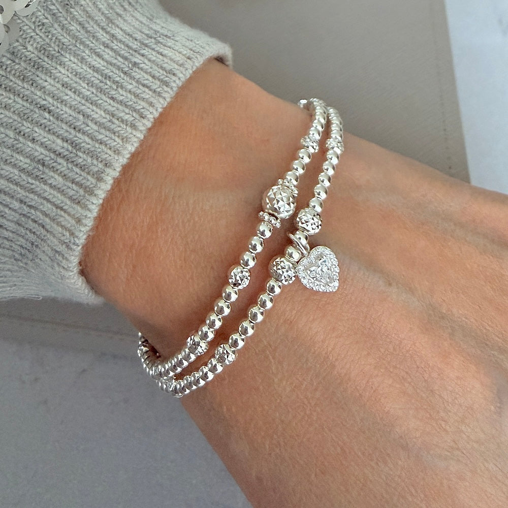 Two silver beaded stacking bracelets, one with a heart charm on a wrist, against a neutral background