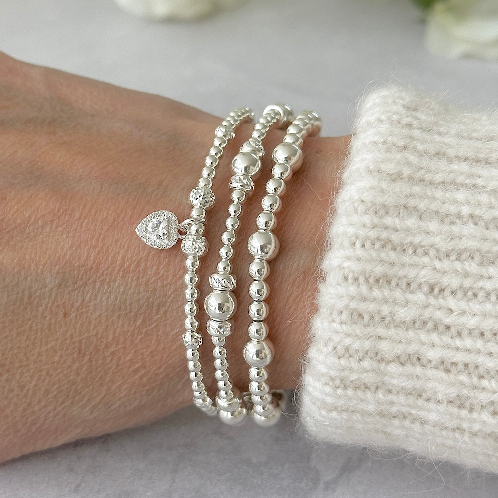 Silver beaded bracelets on a wrist, one bracelet featuring a cubic zirconia heart charm, against a light background