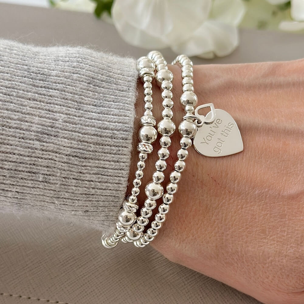 Silver beaded bracelet with a heart-shaped charm with the words 'You've Got This' engraved and a tiny open heart charm sitting on top of it. Worn on a wrist with two other beaded stacking bracelets, against a neutral background.