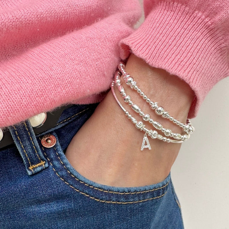 Silver beaded bracelet with letter 'A' charm on wrist with two other silver bracelets, wearing pink sweater and blue jeans.