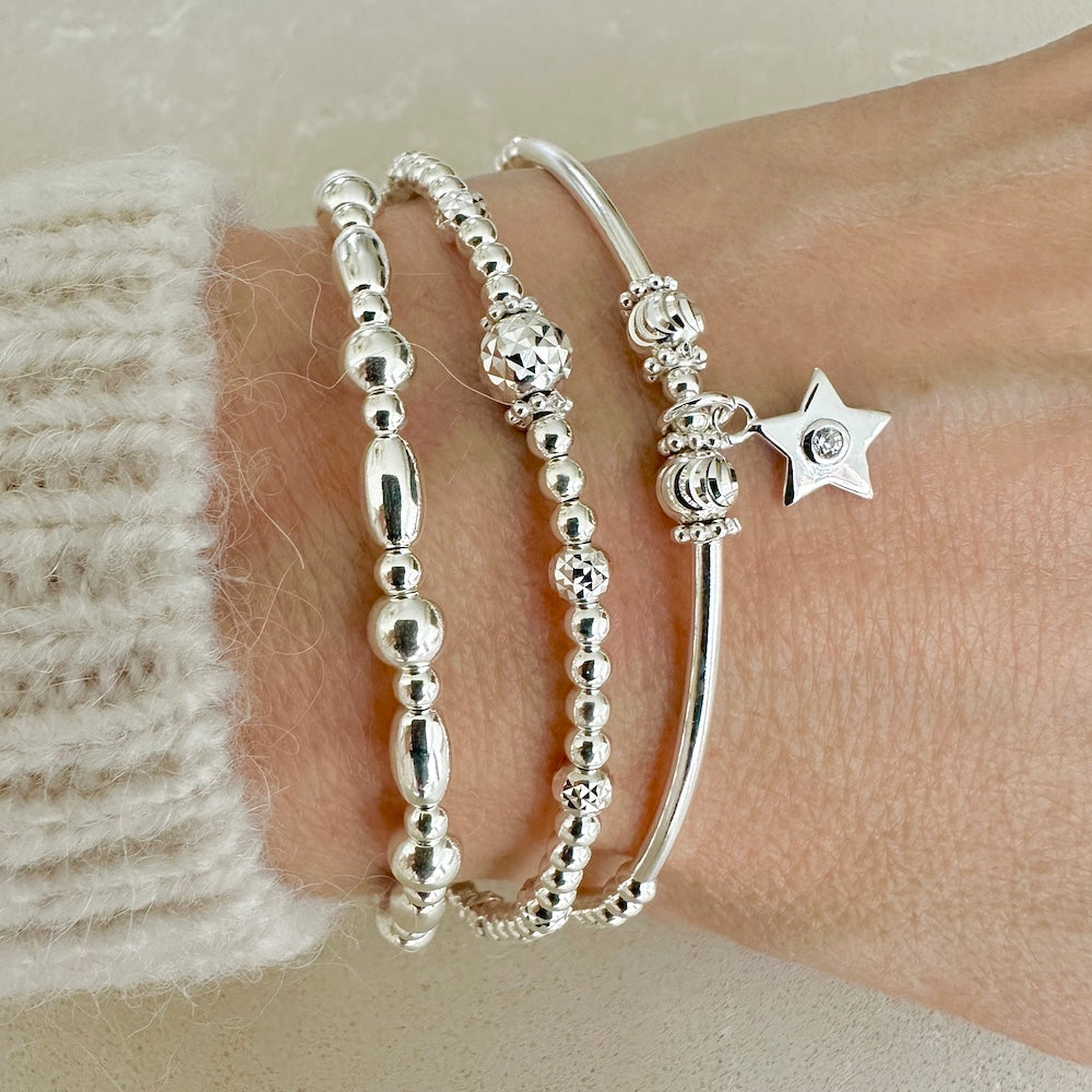 3 silver beaded stacking bracelets, one featuring a small star charm with a tiny cubic zirconia stone in centre of the charm, on a wrist against a neutral background