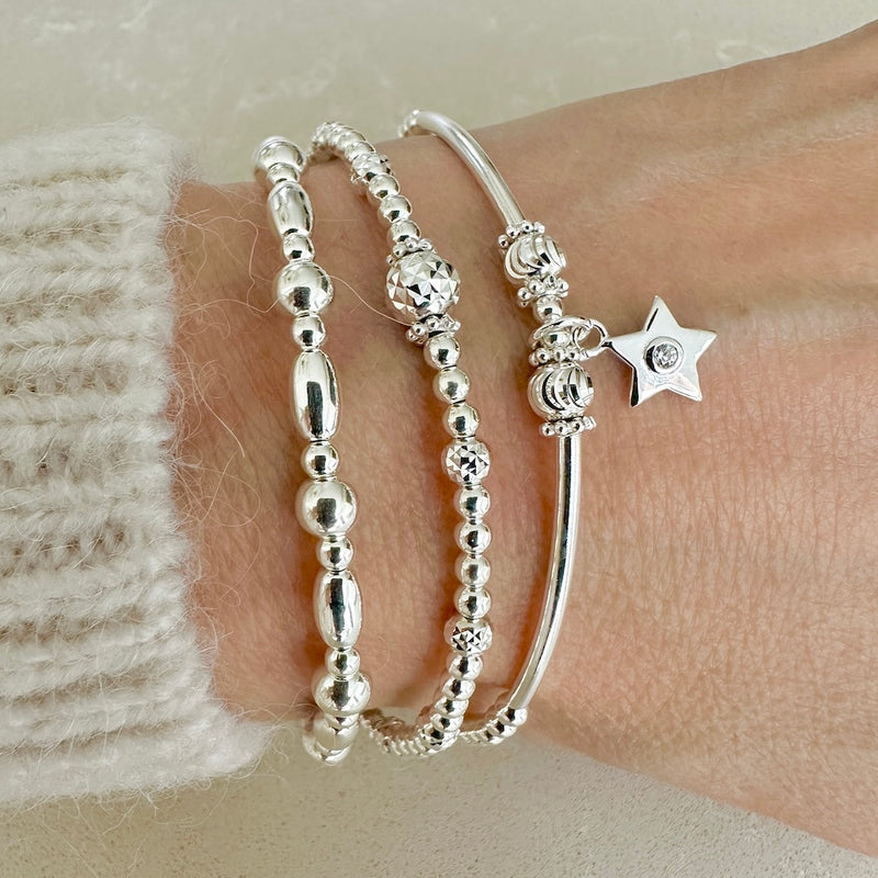 3 silver beaded stacking bracelets, one featuring a small star charm with a tiny cubic zirconia stone in centre of the charm, on a wrist against a neutral background