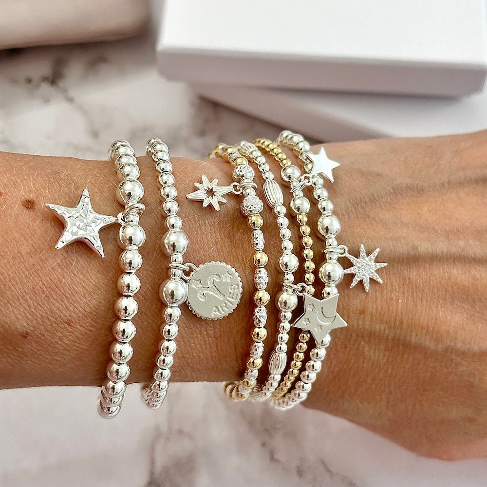 Multiple sterling silver bracelets with star and zodiac charms on a wrist against a marble background
